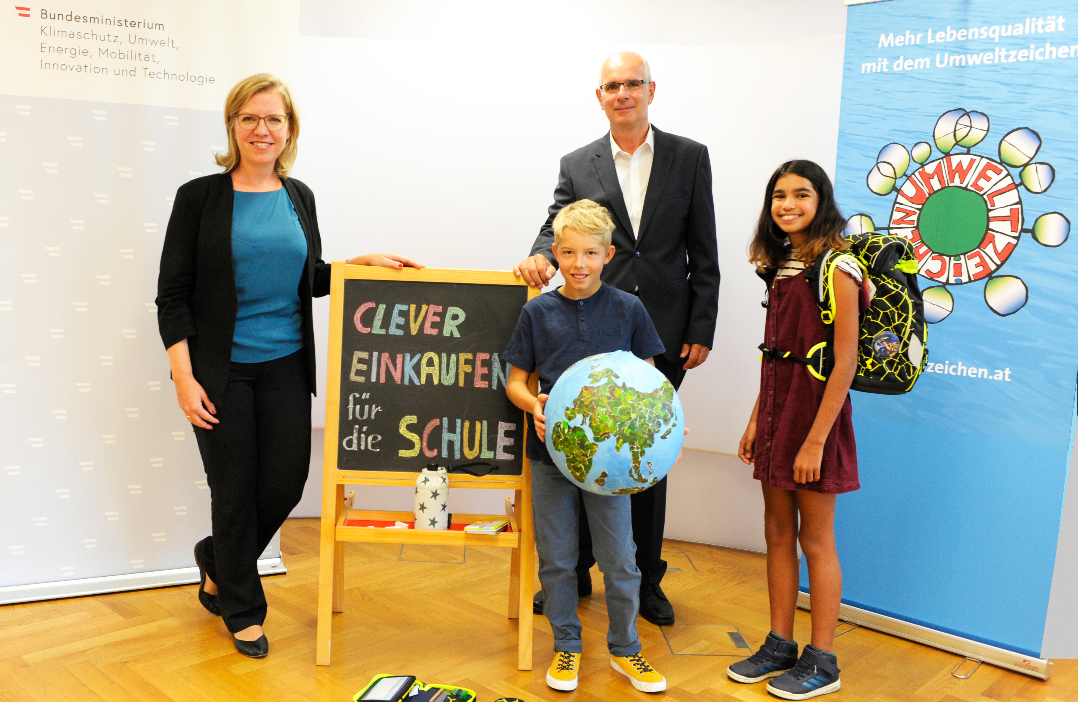 Ministerin Leonore Gewessler mit zwei Schulkindern und einer Tafel mit der Aufschrift "Clever einkaufen für die Schule"